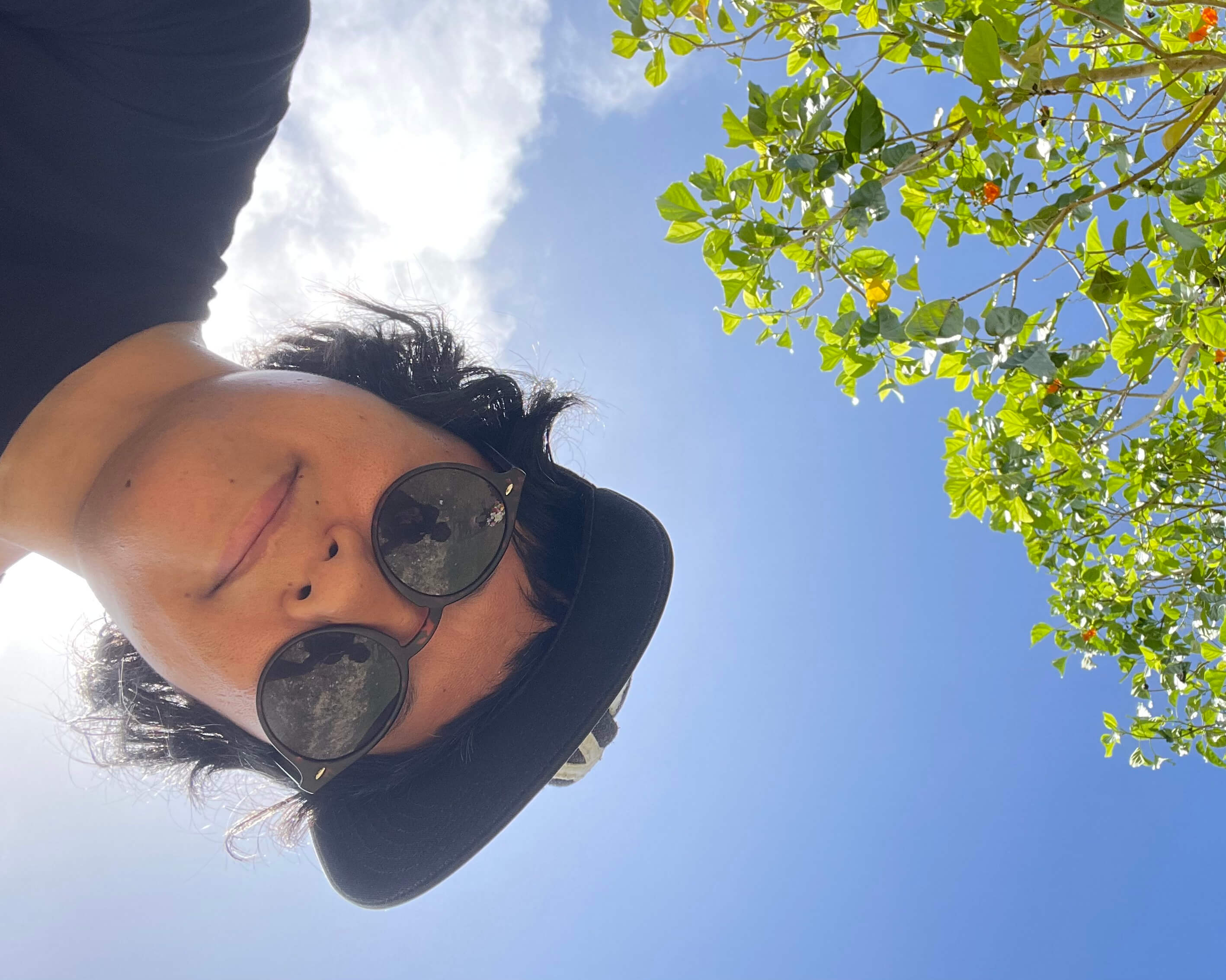 A brown nonbinary person wearing a black t-shirt, sunglasses, and baseball cap looking down at the camera and backlit by sunlight, a cloud, some tree branches, and the sky.